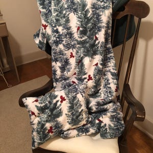 May include: A cozy blanket and pillow set featuring a winter forest design with evergreen trees in shades of blue and gray, accented with red cardinals. The blanket and pillow are draped over a wooden rocking chair.