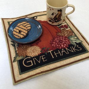 Op de afbeelding: Een blauwe en bruine stoffen placemat met een pompoenontwerp en de tekst "Give Thanks". Een blauwe schotel met twee chocoladekoekjes staat op de placemat. Een witte mok met een herfstscène en een handvat staat op de placemat.