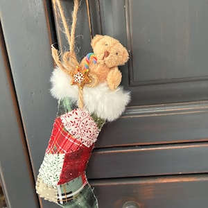 May include: A patchwork Christmas stocking featuring a variety of red, green, and white fabric patterns. The top is trimmed with white faux fur and decorated with a small tan teddy bear, a miniature rainbow candy cane, and a gold star ornament.