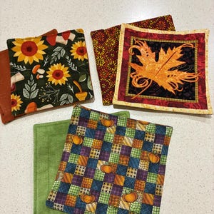 Puede incluir: Un juego de cuatro posavasos cuadrados de tela con varios estampados, incluyendo girasoles amarillos sobre fondo oscuro, una hoja de arce naranja sobre un cuadrado negro, un pequeño estampado floral y una cuadrícula multicolor con calabazas.
