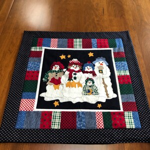 May include: A blue and white quilted table runner with a patchwork design featuring a group of snowmen with stars and a white background. The snowmen are wearing hats and scarves.