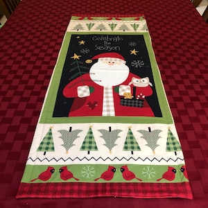 May include: A red, green, and white Christmas table runner with a Santa Claus, an owl, and a Christmas tree. The text "Celebrate the Season" is printed on the runner.