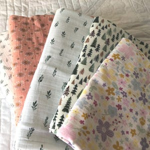 Pode incluir: Cinco mantas de musselina dobradas com vários padrões, incluindo uma estampa floral cor de pêssego, um tecido branco com pequenas folhas verdes, um pano branco com pinheiros verde-escuros e uma manta branca com desenhos florais coloridos.