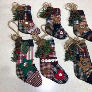May include: Six Christmas stockings with patchwork patterns in red, green, and brown. Each stocking has a jute loop for hanging, and is decorated with a star, button, or heart. Greenery and berries add festive detail.