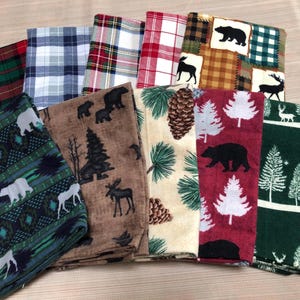May include: A collection of folded fabric napkins with various nature-themed patterns. Designs include plaid, bears, moose, pine cones, and trees in colors like red, green, brown, and cream. These napkins are suitable for rustic or cabin-themed decor.