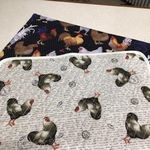 May include: Two rectangular quilted fabric mats. The top mat features a black background with a colorful rooster pattern. The bottom mat has a light cream background with a repeating rooster print, cursive script text, and white trim edges.