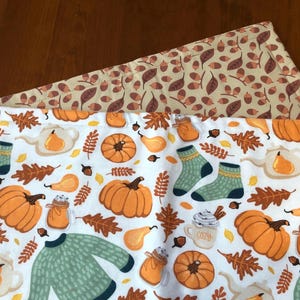 May include: Two rectangular placemats. The top one features a white background with a repeating pattern of pumpkins, pears, leaves, socks, and a sweater. The bottom placemat has a tan background with a pattern of leaves and acorns.