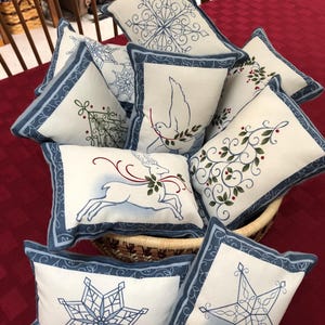 May include: A collection of eight decorative throw pillows with blue trim and various embroidered designs, including snowflakes, a dove, a deer, and a star. The pillows are arranged in a wicker basket on a red checkered tablecloth.