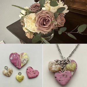 May include: A bouquet of roses and peonies in shades of mauve and cream, with green foliage, sits in a clear glass vase. Below, heart-shaped pendants and a necklace with heart charms in pink, yellow, and white are displayed.