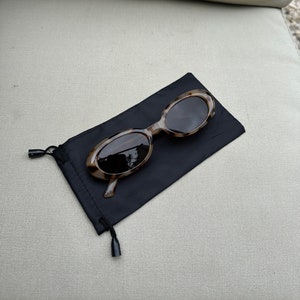 May include: Oval-shaped sunglasses with a tortoiseshell pattern rest on a black drawstring pouch. The sunglasses have dark lenses and a light brown and dark brown frame. The pouch is made of a soft, matte material.