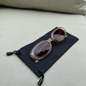 Puede incluir: Gafas de sol ovaladas con montura marrón claro y lentes oscuras sobre una bolsa negra con cordón. Las gafas tienen una montura transparente y un diseño clásico. La bolsa está hecha de un material suave y mate.