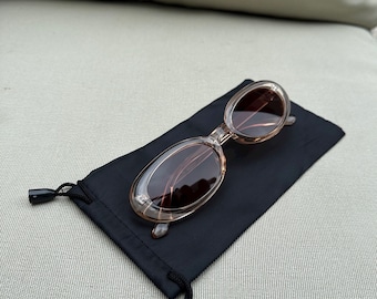 May include: Oval-shaped sunglasses with a light brown frame and dark lenses rest on a black drawstring pouch. The sunglasses have a transparent frame and are designed in a classic style. The pouch is made of a soft, matte material.
