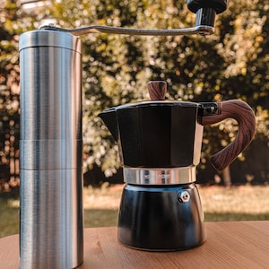 Puede incluir: Un molinillo de café manual de plata con una cafetera moka negra y marrón sobre una superficie de madera. El molinillo tiene una manivela y un recipiente transparente para granos de café. La cafetera moka tiene un mango de madera y un cuerpo de metal negro.