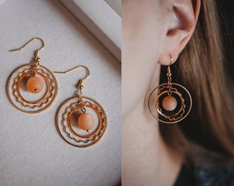 May include: Gold hoop earrings with a peach-colored glass bead in the center. The earrings have a delicate, layered design.