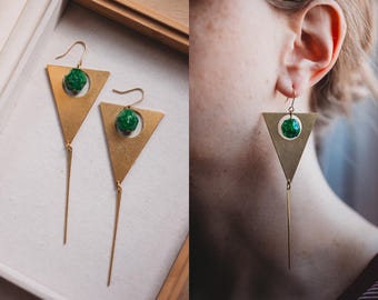 Peut inclure: Une paire de boucles d'oreilles en forme de triangle dorées avec un pendentif en pierre précieuse verte. Les boucles d'oreilles ont une longue chaîne dorée fine attachée au bas du triangle.