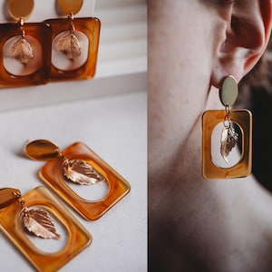 Puede incluir: Un par de pendientes de declaración de oro y ámbar con un diseño de hoja. Los pendientes son de forma cuadrada con un dije de hoja de oro que cuelga de la parte inferior.