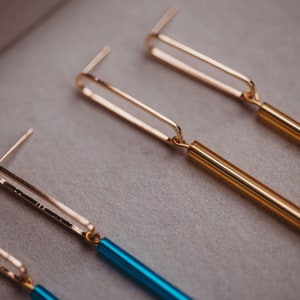 Peut inclure: Deux paires de boucles d'oreilles dor&eacute;es et bleues. Les boucles d'oreilles ont un design simple de barre dor&eacute;e avec une longue barre fine de couleur suspendue au bas. Une paire a une barre dor&eacute;e et une barre dor&eacute;e, tandis que l'autre paire a une barre dor&eacute;e et une barre bleue.