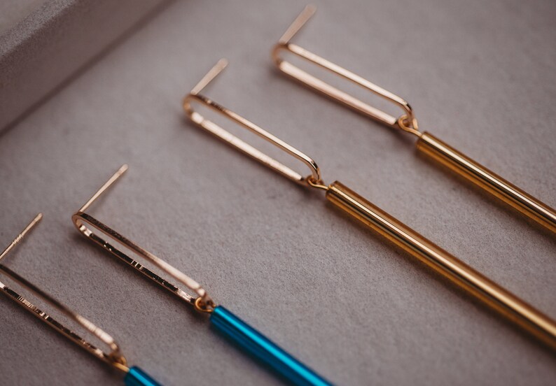 Peut inclure: Deux paires de boucles d'oreilles dor&eacute;es et bleues. Les boucles d'oreilles ont un design simple de barre dor&eacute;e avec une longue barre fine de couleur suspendue au bas. Une paire a une barre dor&eacute;e et une barre dor&eacute;e, tandis que l'autre paire a une barre dor&eacute;e et une barre bleue.