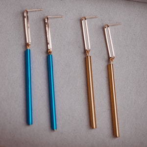 Peut inclure: Quatre paires de boucles d'oreilles pendantes avec des anneaux dor&eacute;s et de longues tiges fines et color&eacute;es. Deux paires ont des tiges bleues et deux paires ont des tiges dor&eacute;es.