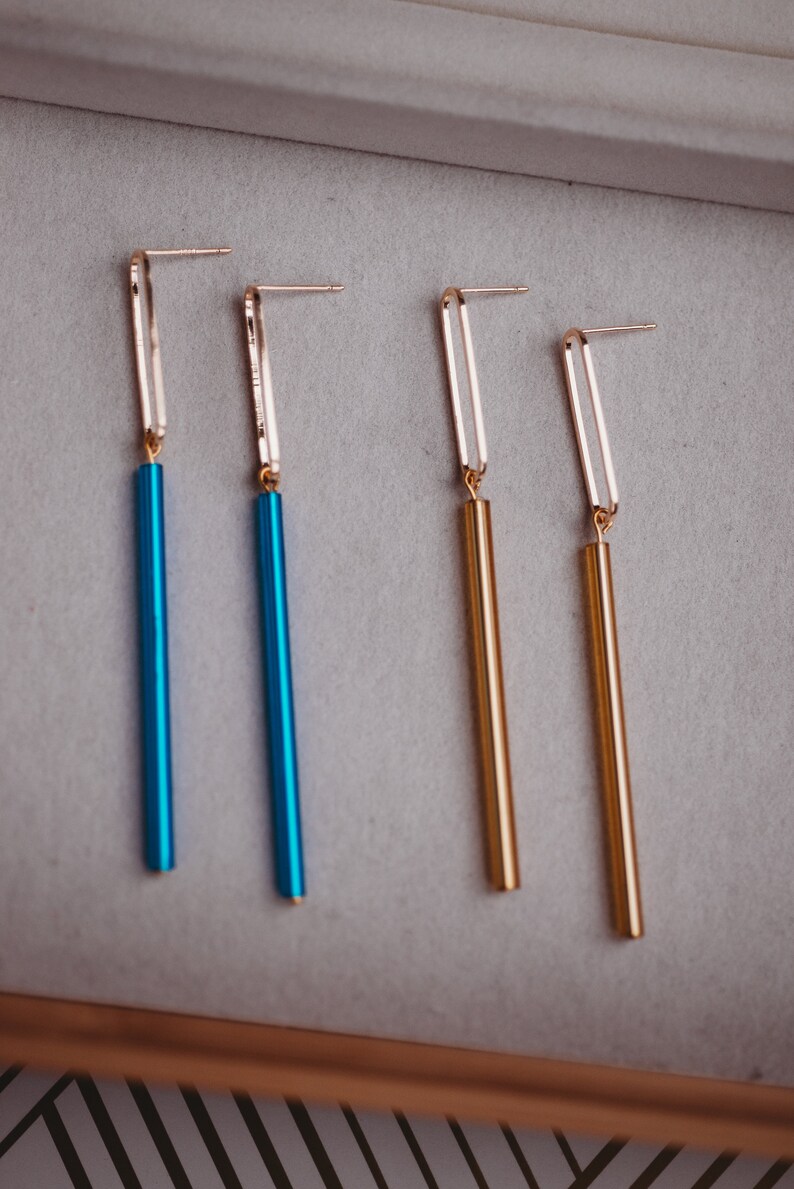 Peut inclure: Quatre paires de boucles d'oreilles pendantes avec des anneaux dor&eacute;s et de longues tiges fines et color&eacute;es. Deux paires ont des tiges bleues et deux paires ont des tiges dor&eacute;es.