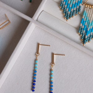 May include: A pair of gold beaded earrings with a gradient of blue and turquoise beads. The earrings are displayed in a white jewelry box.
