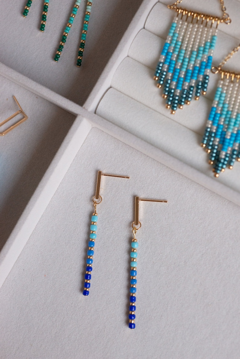 May include: A pair of gold beaded earrings with a gradient of blue and turquoise beads. The earrings are displayed in a white jewelry box.