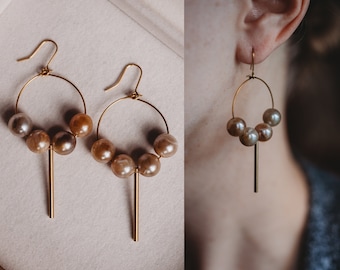 May include: A pair of gold hoop earrings with a dangling bar and five iridescent brown pearls. The earrings are shown on a white surface and on a person's ear.