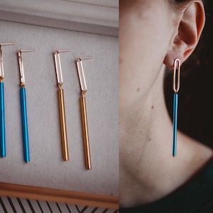 May include: Four pairs of long, thin dangle earrings. Two pairs are gold and two pairs are blue. The earrings are shown on a white surface and on a person's ear.
