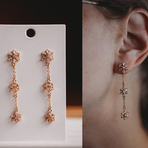 May include: Rose gold drop earrings with a snowflake design. Each earring features three snowflake-shaped charms linked by fine chains. The earrings are presented on a white card.