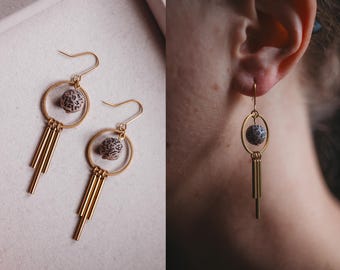 May include: A pair of gold earrings with a small, round, speckled stone in the center of each earring. The earrings have a dangling design with multiple gold bars hanging from the bottom of each earring.