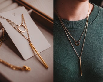 Puede incluir: Collar dorado de varias capas con una cadena fina, un pequeño colgante circular y un colgante de barra vertical más largo. El collar se muestra en una tarjeta blanca y también se usa, sobre un fondo verde.