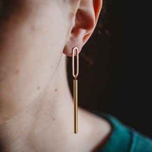 Peut inclure: Boucles d'oreilles pendantes dor&eacute;es avec une longue barre fine et une petite boucle ovale en haut. Les boucles d'oreilles sont port&eacute;es par une personne portant des lunettes.