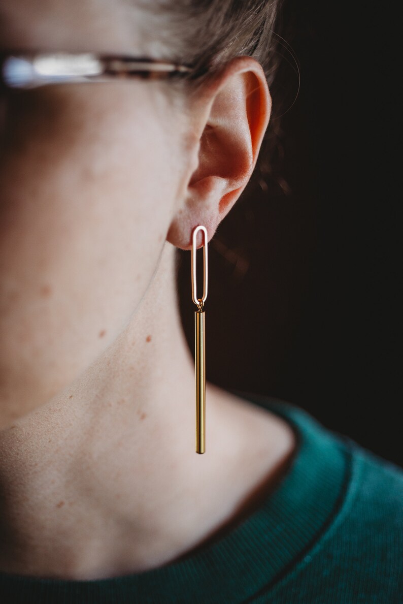 Peut inclure: Boucles d'oreilles pendantes dor&eacute;es avec une longue barre fine et une petite boucle ovale en haut. Les boucles d'oreilles sont port&eacute;es par une personne portant des lunettes.
