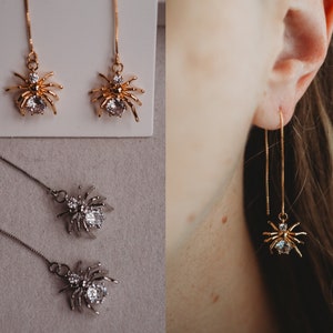 Puede incluir: Un par de pendientes de araña dorados con detalles de cristal transparente. Los pendientes son colgantes y tienen una cadena larga.