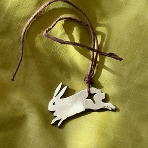 May include: A silver-toned metal pendant shaped like a leaping rabbit with a star-shaped cutout near the tail. The pendant is suspended from a thin, dark brown cord that is knotted through a hole in the upper body of the rabbit figure.