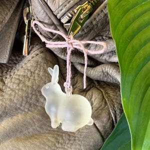 May include: A frosted glass bunny charm is tied to a gray leather bag with pink string. The bunny has long ears and a round body. A gold zipper is visible.