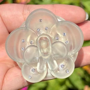 May include: A translucent, pale, off-white flower-shaped accessory featuring a smooth, rounded petal design. The center and petals are accented with small, embedded clear crystals that catch the light, creating a subtle, shimmering effect.