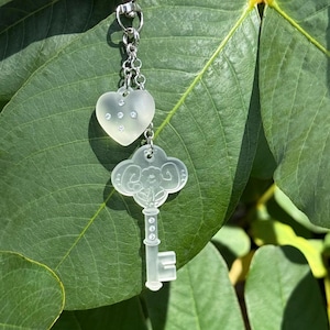 May include: A clear, heart-shaped charm with embedded rhinestones hangs from a silver chain, connected to a key-shaped charm. The key charm has a floral design and is also clear. The charms are set against a backdrop of green leaves.