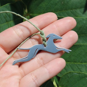 May include: A silver-toned metal pendant shaped like a running greyhound dog, featuring a small star-shaped cutout near the neck. The pendant is suspended from a thin, light green cord that is knotted through the cutout in the metal figure.