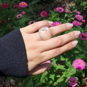 May include: A translucent, rounded ring featuring two small, clear circular accents and one tiny red dot embedded on the surface. The ring is worn on a finger, showcasing its smooth, matte texture and minimalist, rounded shape against a natural backdrop.