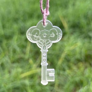 May include: A clear, decorative key pendant with a floral design and a pink string for hanging. The key's head features a four-leaf clover shape with a flower in the center. The key is hanging against a blurred green background.