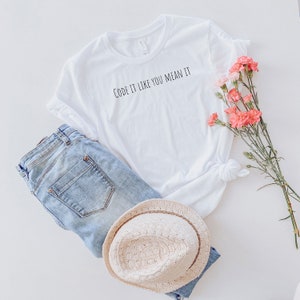 Può includere: T-shirt bianco con la scritta nera "CODE IT LIKE YOU MEAN IT", abbinato a jeans in denim azzurro e un cappello di paglia. Fiori rosa aggiungono un tocco di colore all'outfit casual, creando un'estetica rilassata.