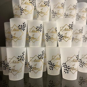 Może przedstawiać: Kolekcja białych kieliszków do wódki. Każdy kieliszek zdobi złoty napis "Adele et Nino" w złotym geometrycznym obramowaniu, ozdobiony czarnymi motywami liści. Kieliszki są ułożone w schludnych rzędach.