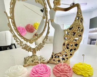 Könnte beinhalten: Vier blütenförmige Kerzen in Weiß, Rosa und Gelb. Die Kerzen sind auf einer weißen Oberfläche vor einem verzierten goldenen Spiegel und einer goldenen und weißen Figur angeordnet.