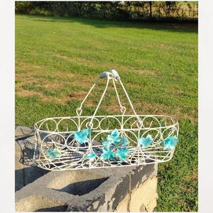 May include: An off-white, oval-shaped metal basket with a handle, adorned with turquoise-colored floral accents. The basket is supported by a concrete block, set against a backdrop of green grass and sunlight.