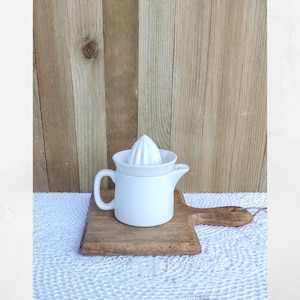May include: A white ceramic citrus juicer featuring a central reamer cone and a pitcher base with a handle and a pouring spout. The juicer is composed of two stackable parts, designed for extracting juice from citrus fruits like lemons or oranges.