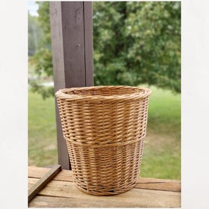 May include: A light brown, woven wicker basket with a cylindrical shape. The basket is made of tightly woven, natural-colored wicker and sits on a wooden surface. The basket is designed for storage or decorative purposes.