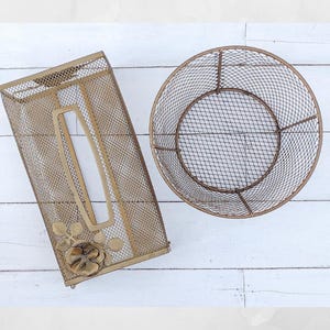 May include: Two decorative metal mesh containers. One is rectangular with a floral accent and a tissue opening. The other is a round basket. Both are in a gold tone and sit on a white wooden surface.