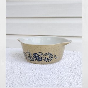 May include: Tan and blue floral patterned bowl. The bowl has a speckled tan exterior and a white interior. The blue floral design wraps around the exterior of the bowl. The bowl has two handles.