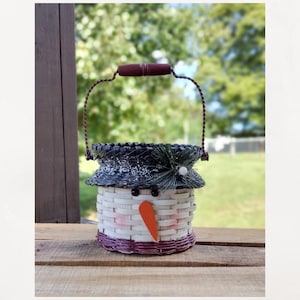 May include: A snowman-shaped basket with a dark brown handle and a woven design. The basket features a black hat with white accents, a carrot nose, and black eyes. The base and rim are a deep purple.